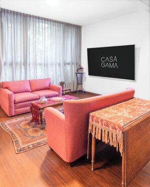 Ein Wohnzimmer mit einem rosa Sofa, einem Couchtisch, einem gemusterten Teppich und einem an der Wand befestigten Flachbildfernseher.