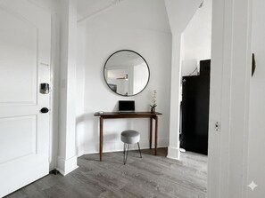 Una habitación con una puerta blanca, un espejo redondo en la pared, un escritorio de madera con una computadora portátil y un taburete gris.