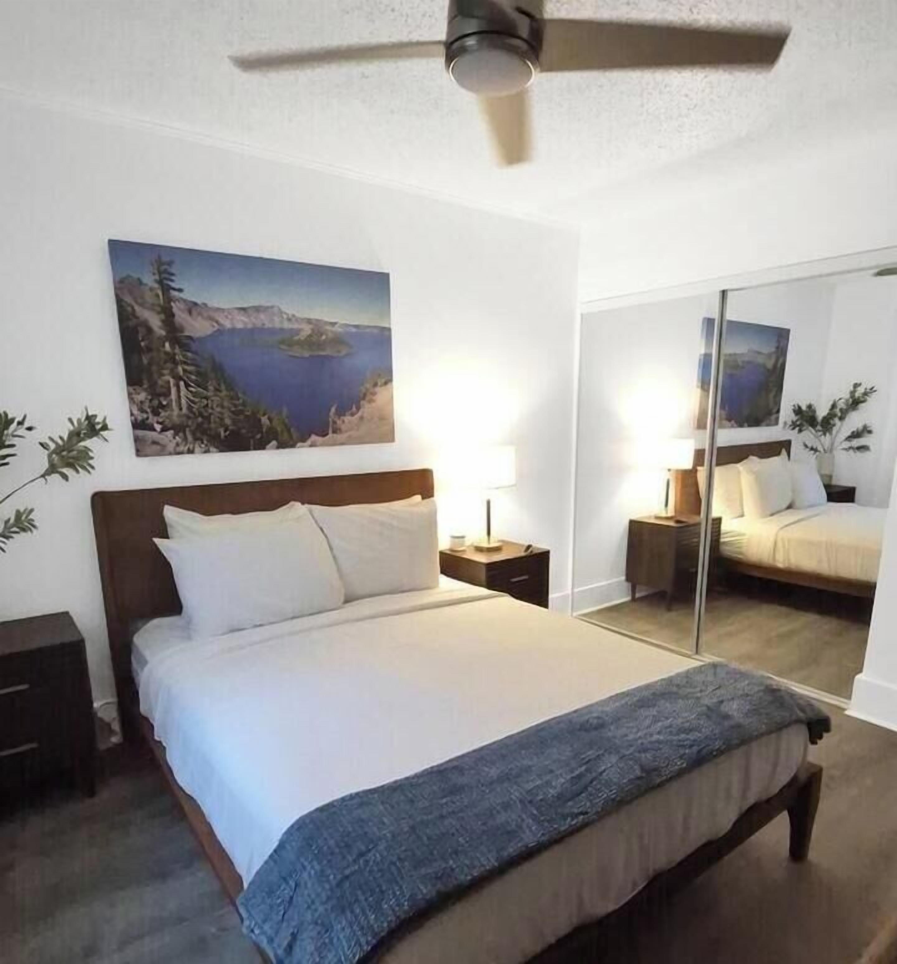 A bedroom with a bed, bedside tables, a ceiling fan, a large mirror, and a painting of a landscape.