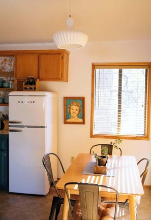 A kitchen with a white refrigerator, wooden cabinets, a dining table with chairs, a hanging pendant light, and a window with blinds. - Idyllwild Cabin w/ Mountain Views & Deck (Idyllwild-Pine Cove)