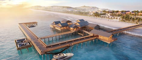 Un complexe hôtelier situé sur pilotis, composé de plusieurs hébergements de type bungalow reliés par des passerelles, et offrant un accès à une plage et à une jetée.