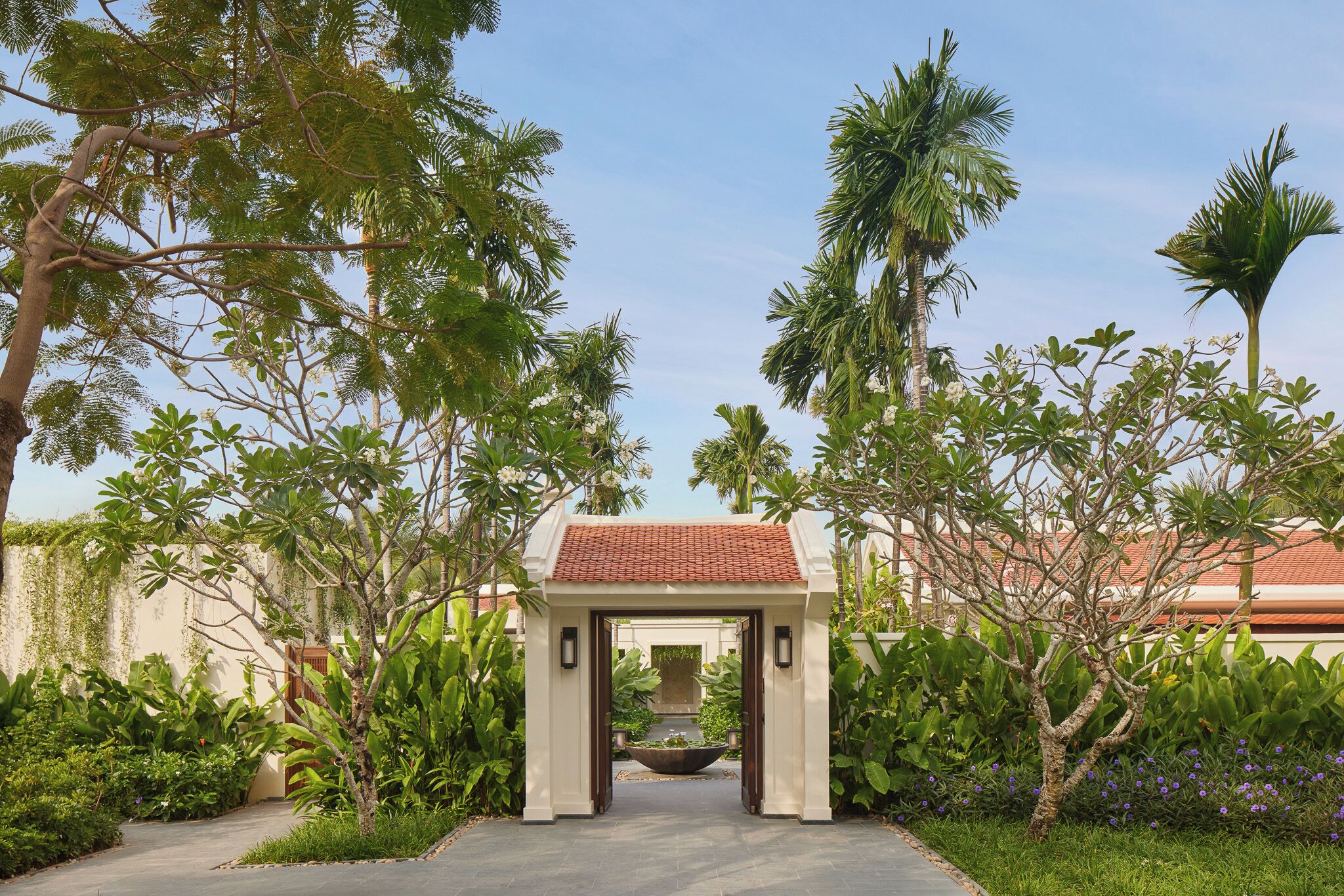A pathway leading to a building with a tiled roof, surrounded by lush greenery and palm trees.