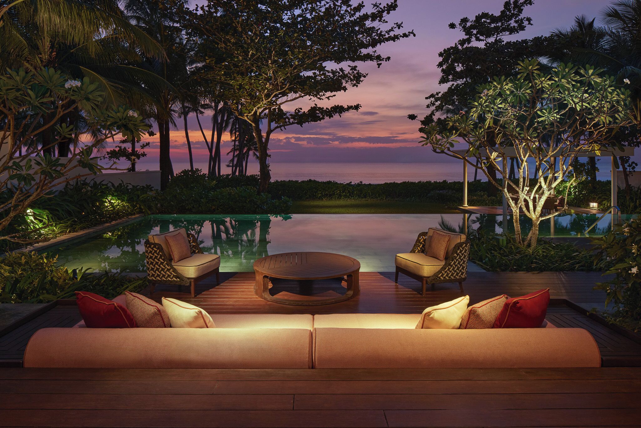An outdoor seating area with a pool, surrounded by tropical vegetation and a sunset view.