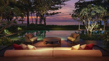 An outdoor seating area with a pool, surrounded by tropical vegetation and a sunset view.