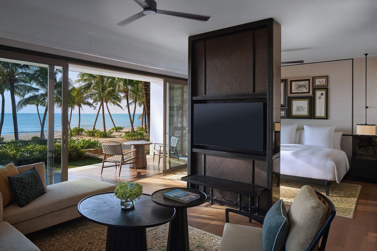 A modern hotel room with a large TV, a balcony with a view of the beach and palm trees, a sofa, and a dining area.