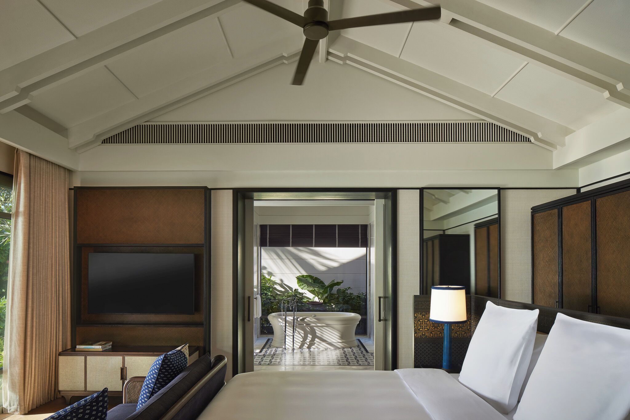 A hotel room with a bed, a TV, a ceiling fan, a bathroom with a bathtub, and a view of the outdoors.