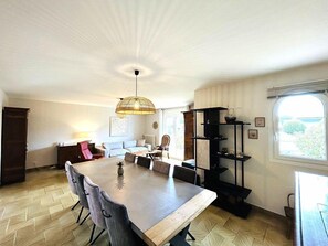 A dining area with a large table, chairs, a hanging light fixture, and a window with a view. - Beautiful family home with garden, terrace, wifi, near Guidel beach (GUIDEL)