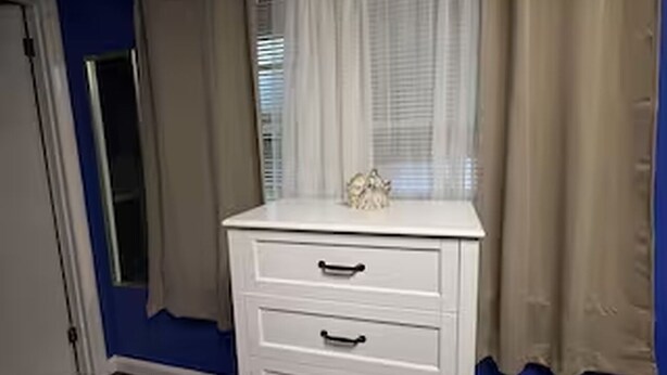 Ein Zimmer mit weißem Schminktisch, einem Fenster mit Vorhängen und einer Tür.
