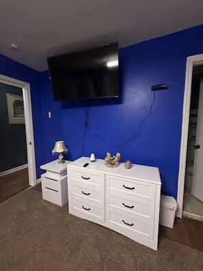 Habitación con pared azul, cómoda blanca con cajones, televisor de pantalla plana montado encima y una lámpara sobre la cómoda. Se ve una puerta a la izquierda.