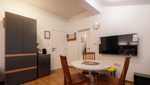 Una cocina compacta con refrigerador, una mesa de comedor con sillas, un televisor montado en la pared y una puerta que da a otra habitación.