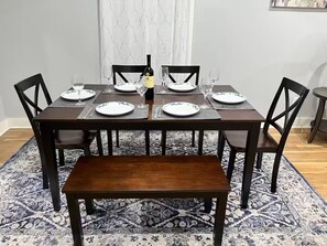A dining table set with plates, glasses, and a bottle, surrounded by chairs and a bench, with a patterned rug and a window with curtains in the background. - Dino's Place - Minutes from Downtown Houston. (Houston)