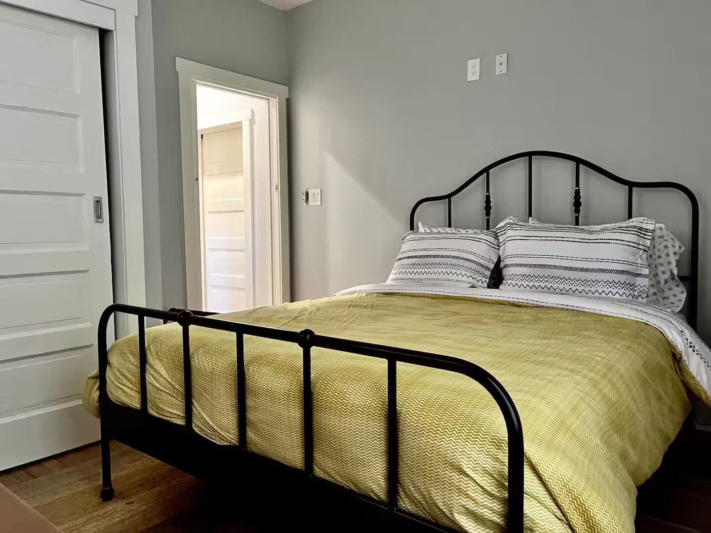 A single bed with a metal frame, a yellow bedspread, and patterned pillows in a room with a white door and light gray walls.