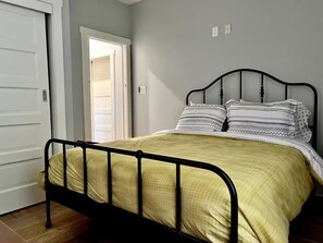 Een enkel bed met een metalen frame, een gele dekbedovertrek en gestreepte kussens in een kamer met een witte deur en lichtgrijze muren.