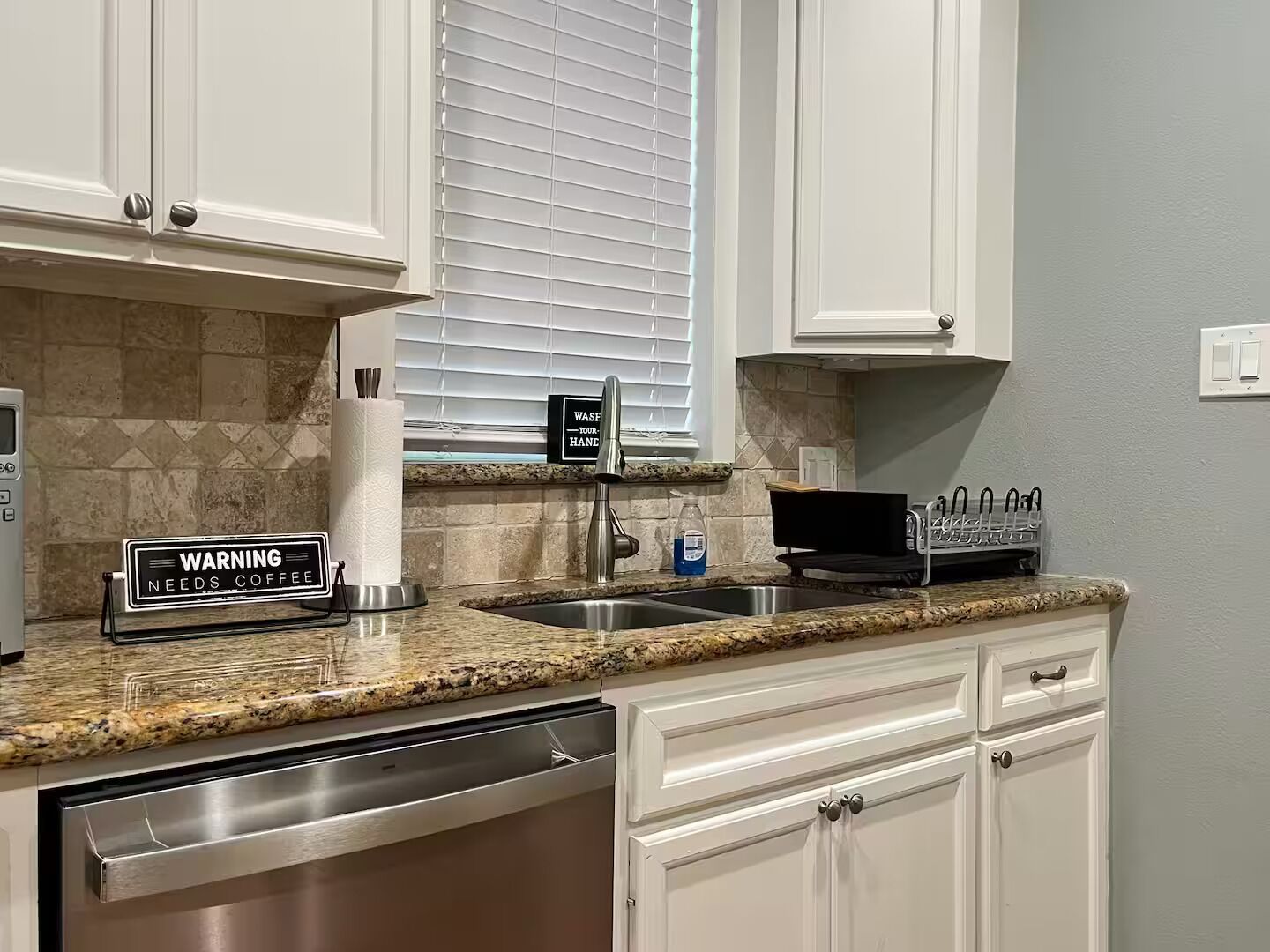 A kitchen with white cabinets, a granite countertop, a stainless steel dishwasher, a sink, and a window with blinds.