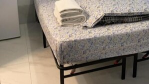 Een enkel bed met een bloemenbedekking, een kussen en een opgerolde handdoek.