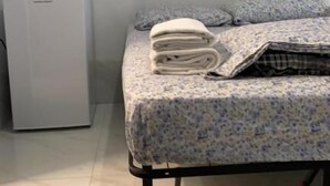 Een bed met een bloemenbedekking en opgerolde handdoeken, een klein airconditioningsapparaat en een aan de muur hangend schilderij.