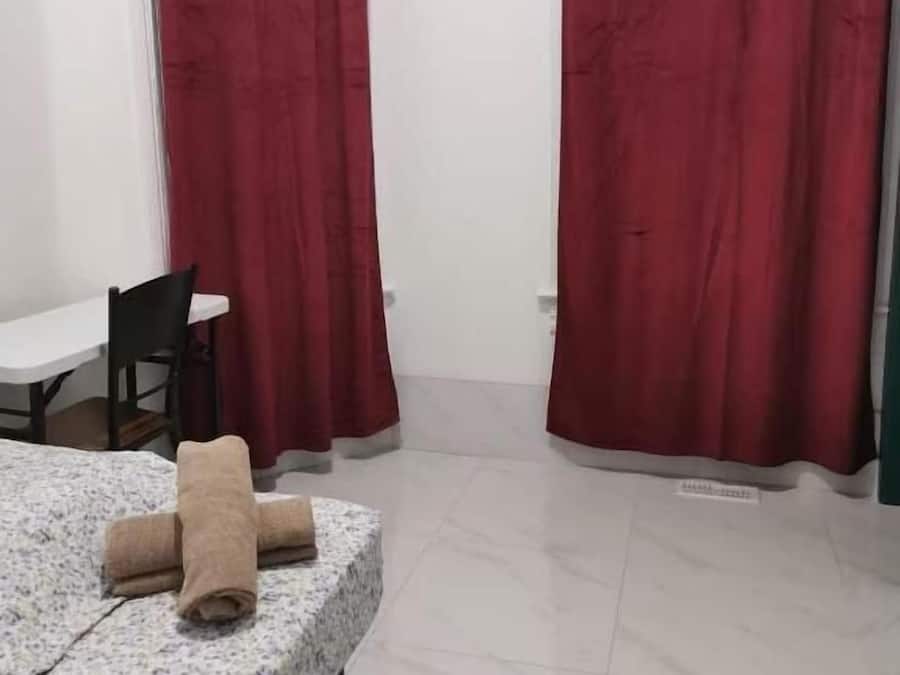 Una habitación con cama, una mesa y una silla. Hay cortinas rojas y un cuadro en la pared.