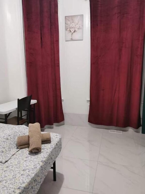 A room with a bed, a table, and a chair. There are red curtains and a framed picture on the wall.