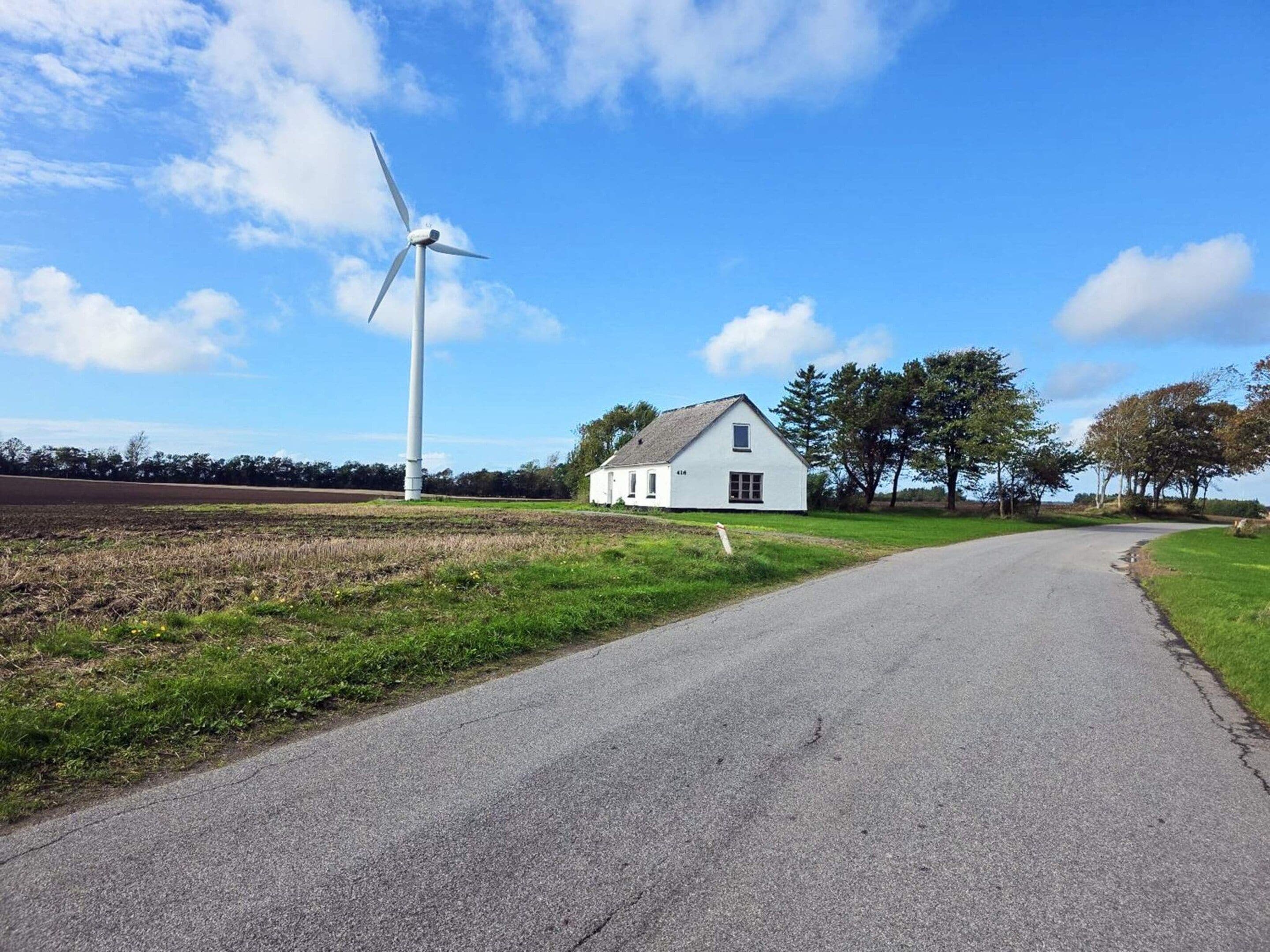 Un camino rural que lleva a una casa blanca, con un aerogenerador al fondo.