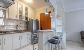 Una cocina moderna con armarios blancos, un refrigerador de acero inoxidable, una estufa a gas y una pequeña barra con dos taburetes.