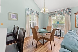 A dining area with a glass-top table surrounded by wicker chairs, a wooden dining table, and a view of the outdoors through large windows. - Cumberland Terrace 2E (Myrtle Beach)