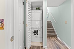 A laundry room with a stacked washer and dryer, a door with a brass handle, and a staircase leading to another level. - Cumberland Terrace 2E (Myrtle Beach)