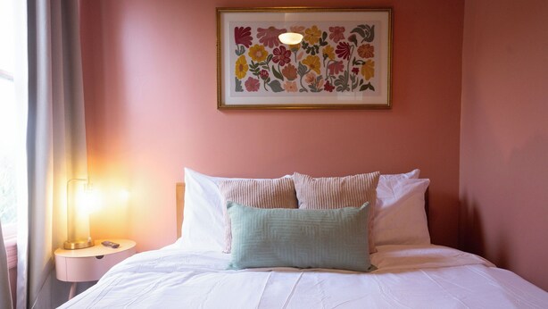 Ein ordentlich bezogenes Bett mit weißen Leinentüchern und zwei Kissen, eine Nachttischlampe und ein gerahmtes Blumenbild an der Wand.