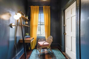 Ein Zimmer mit Holzstuhl, Schreibtisch mit Büchern und Pflanze, Fenster mit gelben Vorhängen und einer Tür.