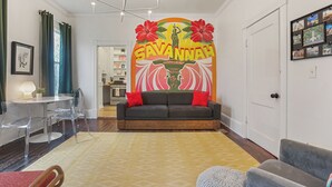 Ein Wohnzimmer mit einer Couch, einem Couchtisch und einem Essbereich mit Stühlen. An der Wand befindet sich ein großes Wandbild und eine Tür mit einem Bilderrahmen.