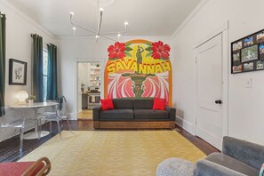 Ein Wohnzimmer mit einer Couch, einem Couchtisch und einem Essbereich mit Stühlen. An der Wand befindet sich ein großes Wandbild und eine Tür mit einem Bilderrahmen.