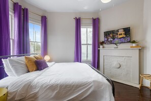 Ein Schlafzimmer mit einem Bett, violetten Vorhängen, einem Gemälde und einer kleinen Ablage mit Pflanzen.