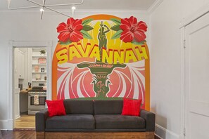 Een woonkamer met een bank en een muurschildering waarop een haan en hibiscusbloemen staan afgebeeld, met de tekst 'SAVANNAH' duidelijk zichtbaar.