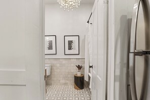 Un baño moderno con suelo de baldosas geométricas, un lavabo blanco, un inodoro y un pequeño taburete de madera con una planta. Hay dos cuadros enmarcados en la pared y una cortina de ducha blanca.