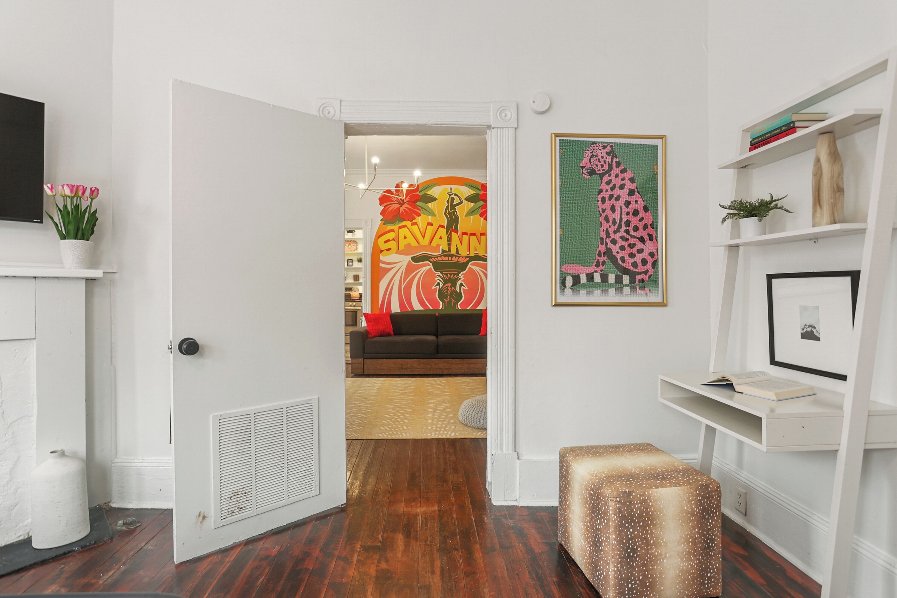 Un pasillo con una puerta blanca que da a una sala de estar. En la pared, se observa una obra de arte enmarcada de un leopardo, un estante blanco con libros y una pequeña planta en maceta.