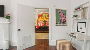 Un pasillo con una puerta blanca que da a una sala de estar. En la pared, se observa una obra de arte enmarcada de un leopardo, un estante blanco con libros y una pequeña planta en maceta.