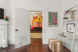 Un pasillo con una puerta blanca que da a una sala de estar. En la pared, se observa una obra de arte enmarcada de un leopardo, un estante blanco con libros y una pequeña planta en maceta.