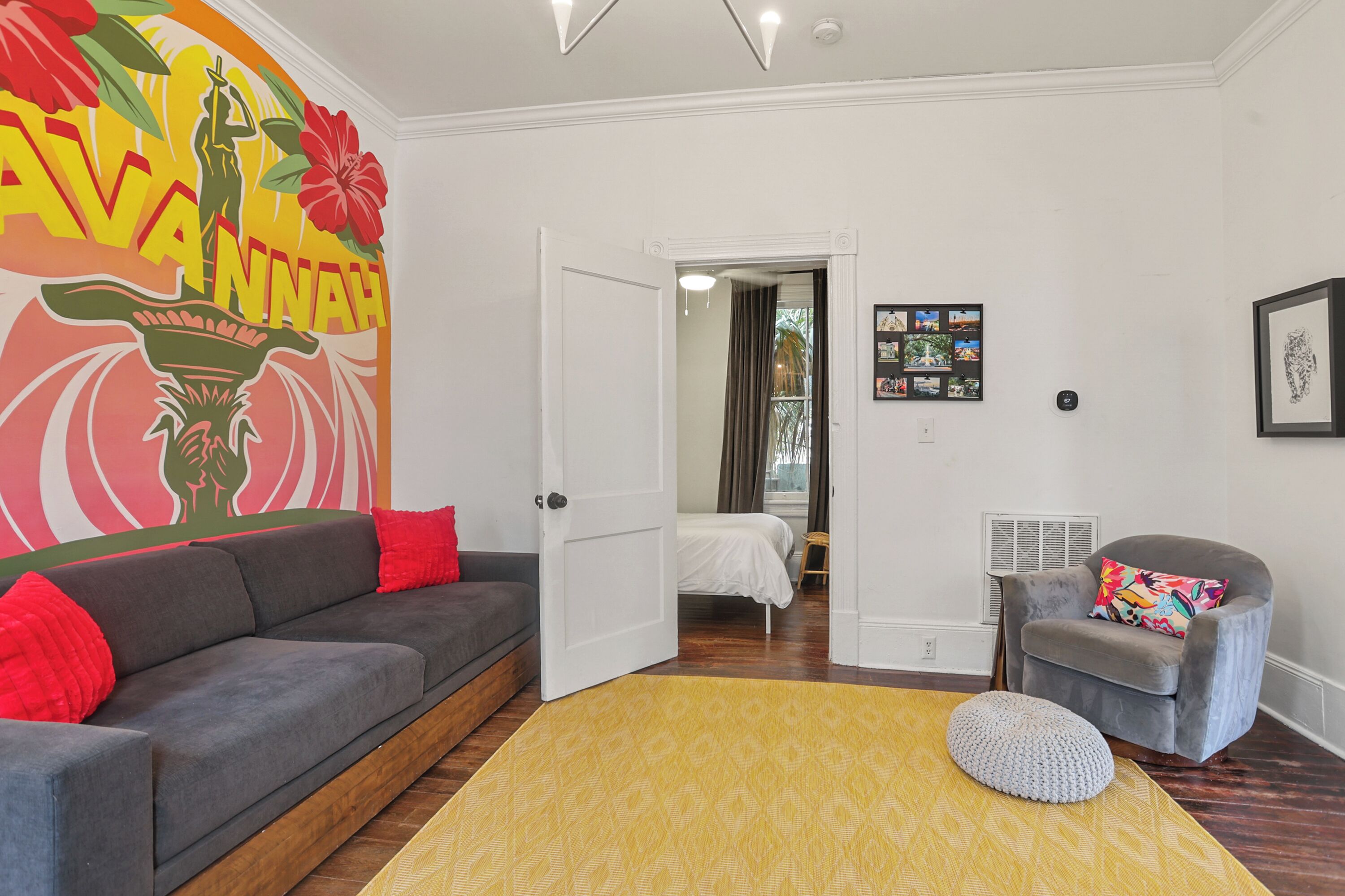 A modern living room with a gray sofa, a yellow rug, and a gray armchair. A white door leads to a bedroom with a bed and a small table.