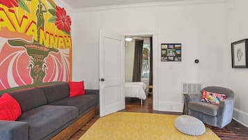 Una sala de estar moderna con un sofá gris, un tapete amarillo y un sillón gris. Una puerta blanca da paso a un dormitorio con una cama y una mesita.