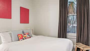 Un lit bien fait, avec du linge de lit blanc et des coussins colorés, une chaise en osier et une fenêtre avec des rideaux.