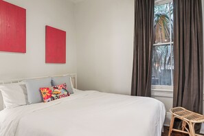 Una cama bien hecha con ropa blanca y almohadas coloridas, una silla de mimbre y una ventana con cortinas.
