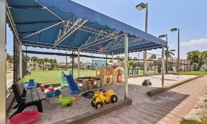 Ein überdachter Spielbereich im Freien mit Sandkasten, Spielgeräten und einem kleinen Gebäude.