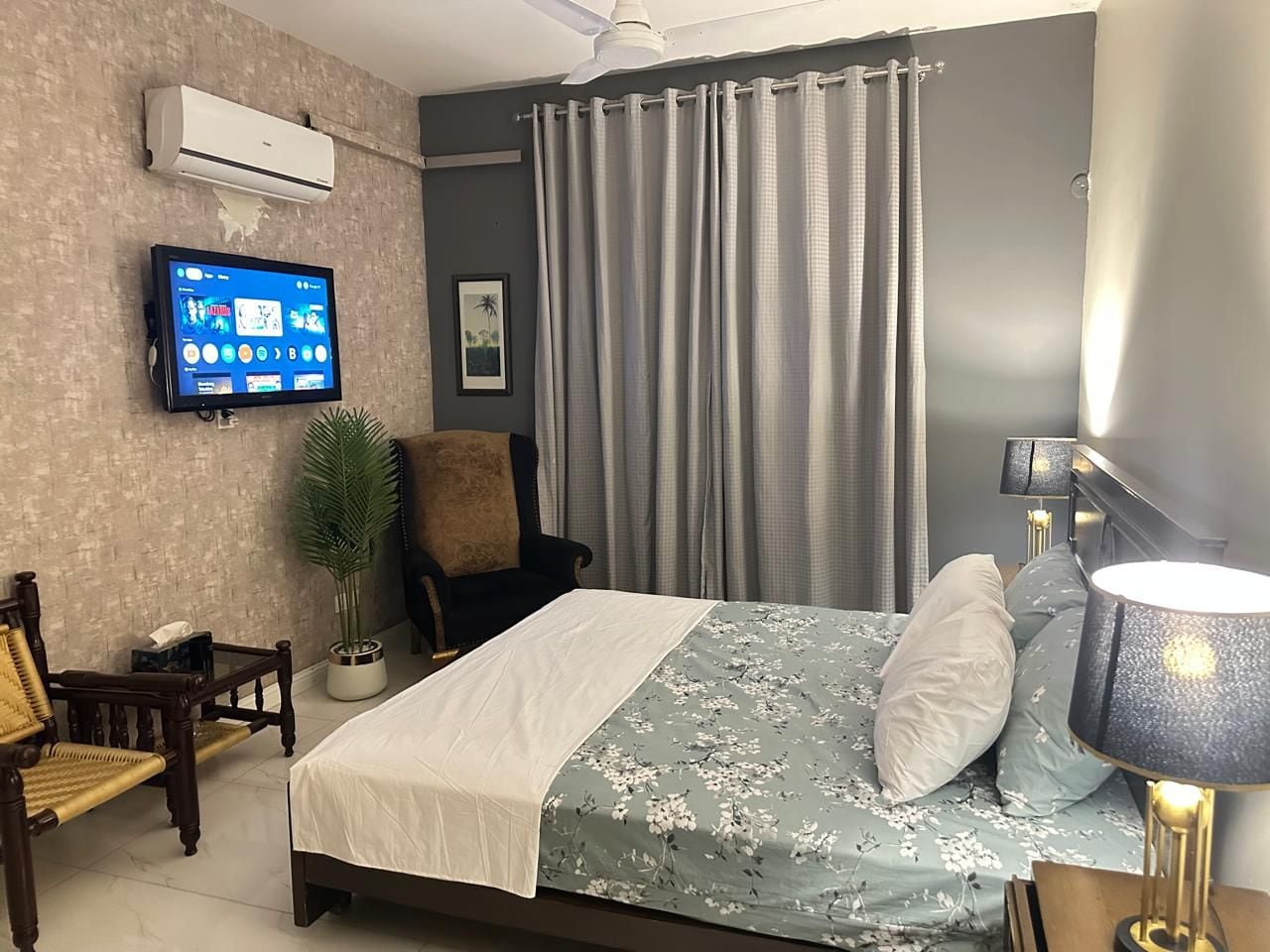 A hotel room with a bed, a television, a chair, a lamp, and a plant.