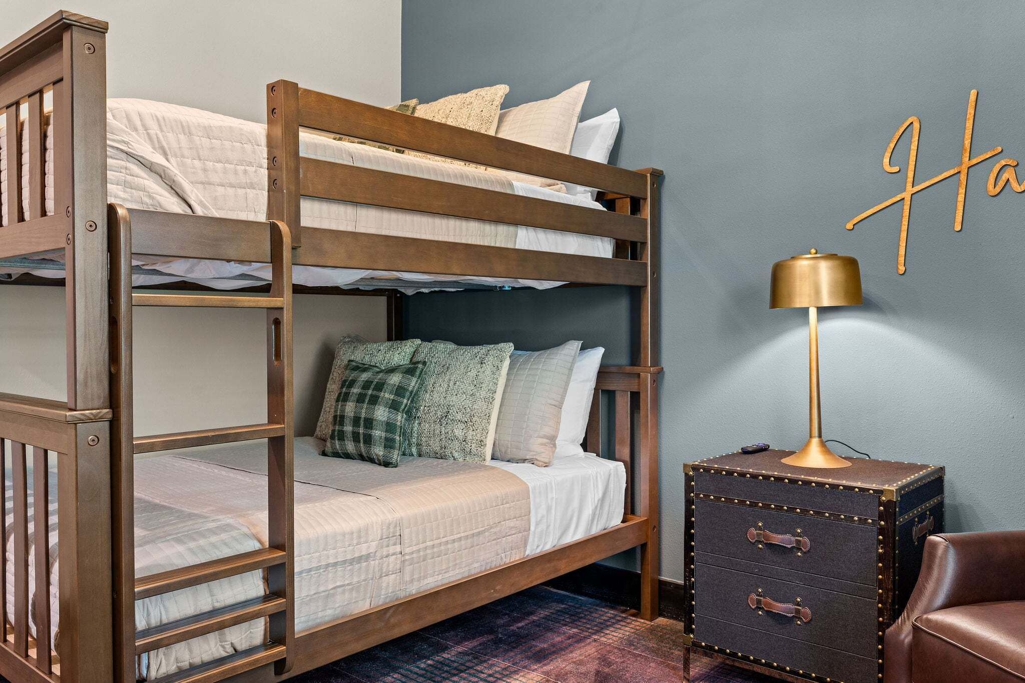 Un lit superposé avec une structure en bois, une table de chevet avec une lampe et un mur orné d’un panneau décoratif.