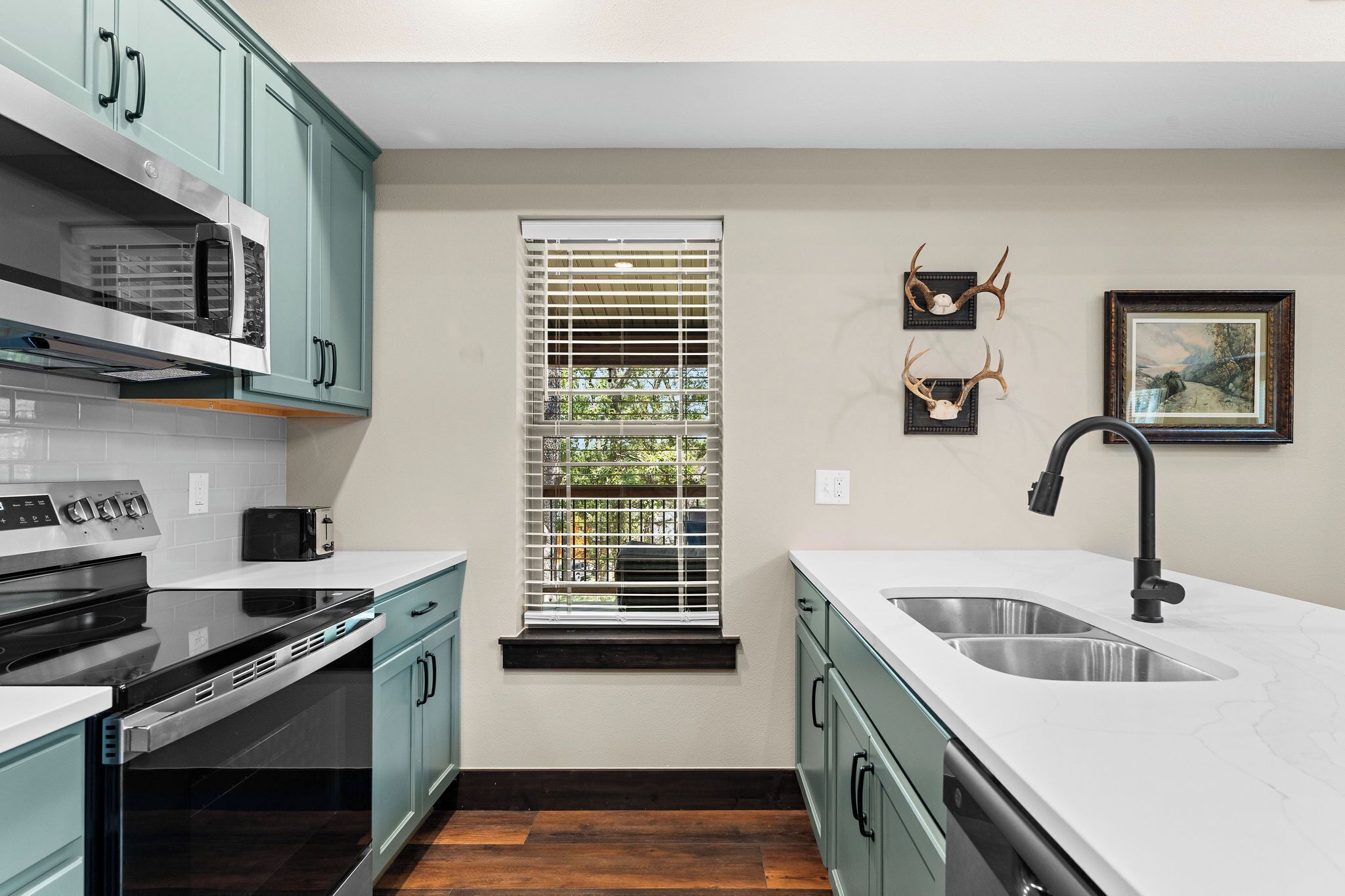 A modern kitchen with teal cabinets, a stainless steel sink, and a window with blinds.