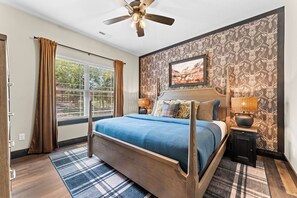Quarto com uma cama grande, um ventilador de teto, uma janela com cortinas, um quadro na parede e um papel de parede estampado.