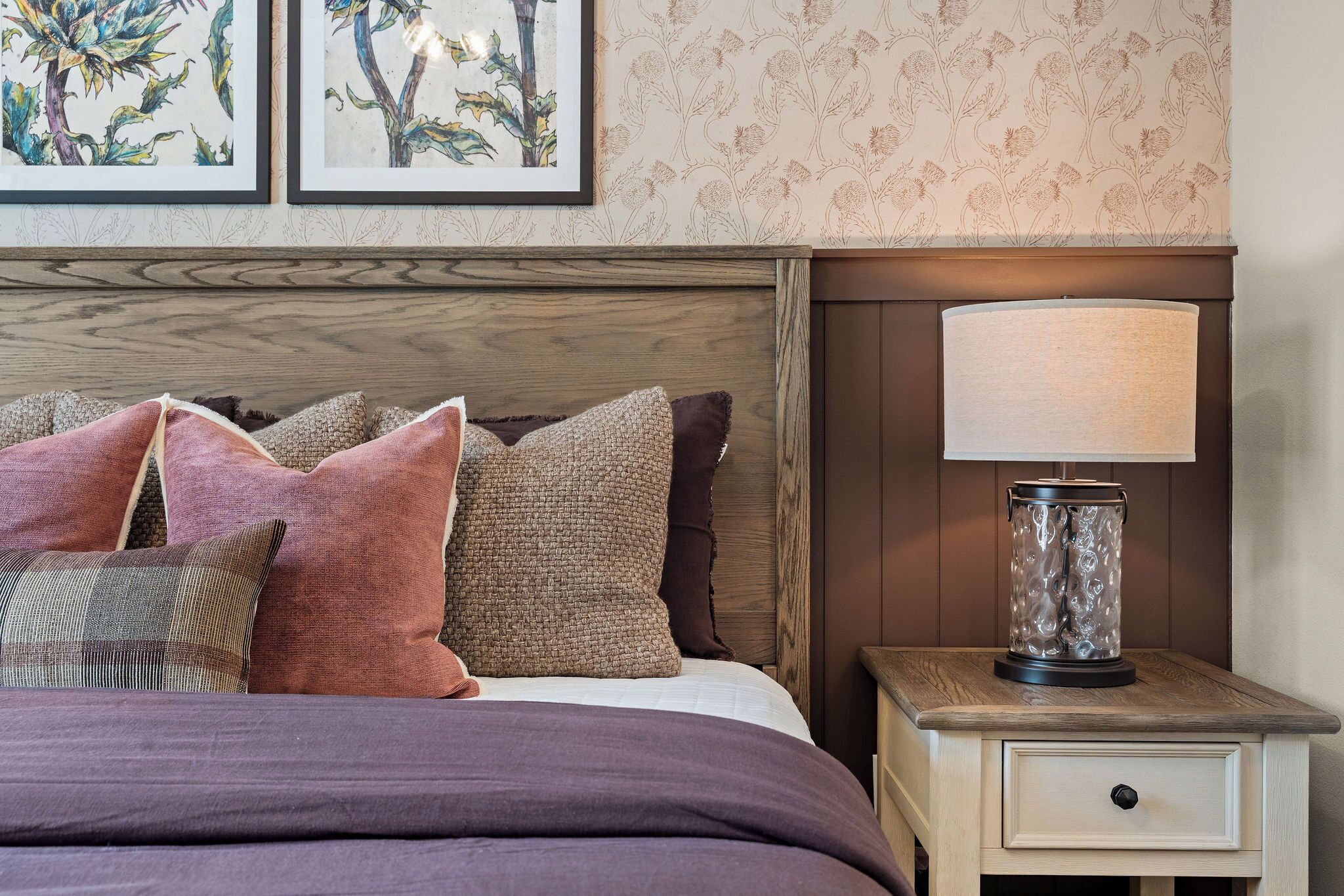 A neatly made bed with a variety of pillows, a bedside lamp, and a wall with floral wallpaper.