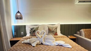 Ein ordentlich bezogenes Bett mit einer braunen, gemusterten Tagesdecke, ein Paar Handtücher, kunstvoll zu einem Schwan geformt, und eine hängende Pendelleuchte.