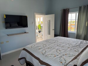 Un dormitorio con cama, televisor montado, puerta que da a un balcón y ventana con cortinas.
