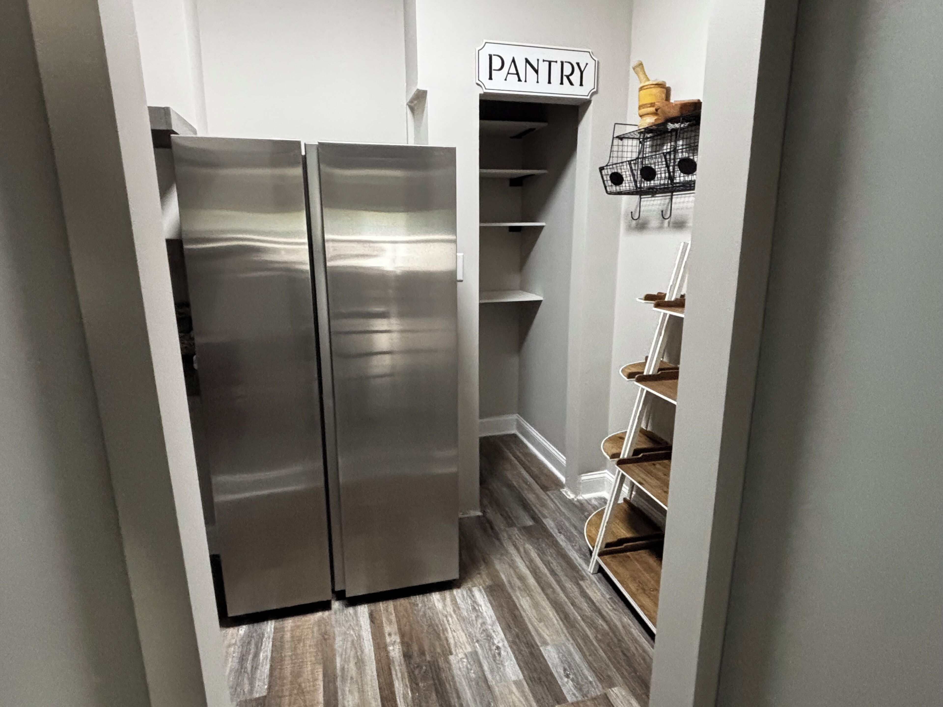 A stainless steel double-door refrigerator in a kitchen with a wooden floor and a pantry area above.