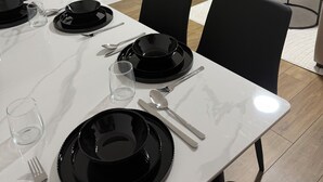 A dining area with a white table set for two, black plates, cutlery, and a glass. A flat-screen TV is mounted on the wall, and a dark-colored sofa is visible in the background.
